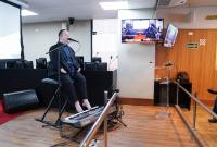 Em um auditório, um homem está sentado diante de um teclado, usando microfone e realizando uma apresentação musical. O ambiente é institucional, com púlpito, telas e piso de madeira ao fundo.