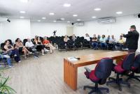 A imagem mostra uma reunião ou palestra em um auditório amplo e bem iluminado. Um homem em pé, à direita, fala ao público, gesticulando enquanto se dirige aos participantes sentados. As pessoas estão distribuídas em cadeiras pretas, dispostas em formato semicircular, acompanhando atentamente a apresentação. À frente, há uma mesa de madeira com cadeiras e um microfone, indicando um ambiente formal de debate ou capacitação. O espaço é organizado, com iluminação moderna.