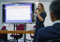Mulher loira em pé ao lado direito, ao lado direito dela, um telão com uma tela branca. Ela está falando com a plateia com um microfone preto na mão esquerda