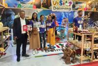 Grupo de pessoas posando para foto, com placas nas maõs. Eles estão em um local repeleto de peças artesanais.