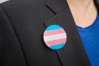 essoa vestindo um blazer preto com um broche redondo preso à lapela, nas cores azul, rosa e branco, associadas à bandeira do orgulho trans.