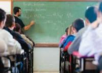 A imagem mostra um professor em sala de aula, escrevendo no quadro enquanto alunos permanecem sentados em suas carteiras, atentos à explicação. O ambiente é escolar, com foco na atividade docente e no processo de ensino-aprendizagem.