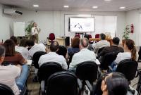 Imagem de uma palestra em uma sala de treinamento. À frente do ambiente, uma mulher em pé fala ao público com um microfone. Ao lado dela, uma tela exibe apresentação sobre a campanha Janeiro Branco, com foco em saúde mental no trabalho. À sua frente, há uma mesa com cadeiras vazias. O público, formado por homens e mulheres adultos, está sentado em fileiras de cadeiras, atento à palestra.