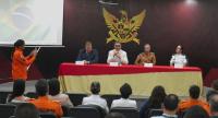 A imagem mostra uma mesa de autoridades em um auditório, com quatro representantes sentados à frente do público, durante um evento institucional. Ao fundo, há um grande brasão oficial na parede, e, à esquerda, uma pessoa em uniforme laranja registra o momento com o celular. A plateia acompanha a cerimônia sentada, em ambiente formal.