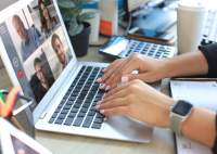 Foto de uma pessoa fazendo teletrabalho. Só aparece as duas mãos dessa pessoa e, na imagem do computador, quatro pessoas em reunião com ela, sendo três homens e uma mulher