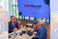 Foto de dois homens de cabelos grisalhos sentados um à frente de outro em um estúdio de rádio. Ambos usam paletó azul marinho
