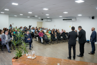 Há três de pé. Eles vestem ternos escuros. Á frente deles há uma público sentado.