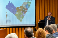 Fotografia de reunião institucional em auditório. Participantes estão sentados assistindo à apresentação de um homem de terno que fala ao microfone. No telão, há um slide com mapa da nova divisão territorial da jurisdição do TRT19, com números de projeção de processos para 2026. O ambiente possui revestimento em madeira clara.   .
