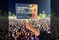 A imagem mostra uma multidão reunida em um grande evento noturno ao ar livre. Em primeiro plano, uma pessoa ergue um cartaz acima da cabeça, chamando a atenção do público. O cartaz traz a mensagem “Viu trabalho infantil? Denuncie! Se calar é permitir”, com referência à Justiça do Trabalho. Ao fundo, milhares de pessoas aparecem concentradas, iluminadas pela luz do evento. A cena transmite um forte caráter de conscientização social em meio a um espaço de grande circulação popular.