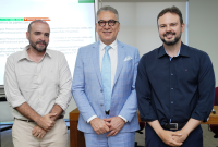 A imagem mostra três homens em pé, lado a lado, posando para a foto em um ambiente interno que aparenta ser uma sala de reunião ou de aula.  Ao centro, um homem de cabelos grisalhos usa óculos e veste terno azul-claro com gravata em tom semelhante, transmitindo formalidade. Ele sorri levemente e mantém as mãos unidas à frente do corpo.  À esquerda, outro homem, com barba curta e cabelos raspados, veste camisa social clara com as mangas dobradas, adotando uma postura mais descontraída.