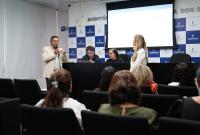 Homem fala ao microfone em auditório institucional, com equipe à mesa ao fundo e público sentado assistindo.