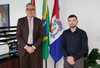 Há dois homens posando para foto. Eles estaõ de pé. O primeiro homem veste terno  escuro e usa óculos. Já o segundo está de camisa de manga comprida, também escura.. Eles estão sorridentes.