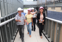 Foto de três mulhers e três homens caminhando em uma passarela de construção. Todos usam roupas informias e capacete branco.