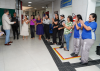 Há um grupo de muleres de pé aplaudindo.. À frente delas há um homem de terno claro. e calça zul. Ele fala para elas.