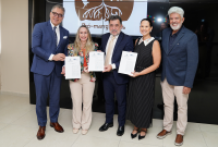 Cinco pessoas em pé, sendo três homens e duas mulheres; dois desses homens estão usando terno escuro e segurando um certificado; as duas mulheres estão usando roupas sociais e também segurando um certificado. O outro homem estão ao lado direito deles, usando uma blazer escuro