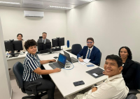 A foto mostra seis pessoas em um ambiente de trabalho, reunidas em uma sala clara com mesas, computadores e notebooks. Parte do grupo está sentada ao redor de uma mesa central com laptops e documentos, enquanto duas pessoas aparecem ao fundo em outra mesa com computadores. Todos estão vestidos de forma profissional e sorrindo para a câmera, sugerindo um momento de trabalho em equipe em uma audiência