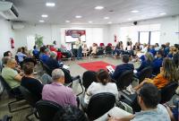 Grupo de pessoas sentadas em um grande círculo. Há um tapete vermelho no centro da sala.