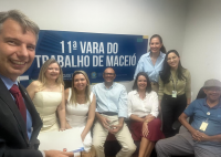 Grupo de pessoas posando para foto. A maioria dela está sentada e há alguma delas em pé. Na parede há escrito 11ª Vara do Trabalho de Maceió. em letras brancas com o fundo azul.