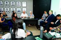 Reunião formal com várias pessoas sentadas; um homem de terno fala ao microfone enquanto os demais escutam.