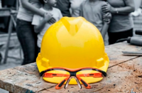 Um capacete de segurança amarelo com óculos de proteção apoiados sobre uma superfície de trabalho, em primeiro plano, enquanto ao fundo aparecem pessoas desfocadas, sugerindo um ambiente de obra ou atividade laboral.