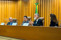 Quatro homens de terno estão sentados atrás de uma mesa em um auditório, participando de uma sessão formal. Ao centro, um deles parece estar falando ou prestando atenção, enquanto os demais mantêm expressão séria. À frente deles há microfones, copos de água e placas com nomes, indicando um evento institucional. Ao fundo, uma cortina dourada compõe o cenário, reforçando o ambiente solene. No primeiro plano, aparecem pessoas sentadas na plateia, observando atentamente a reunião.