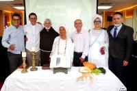 Participantes de diferentes tradições religiosas estão reunidos em um auditório, durante um ato ecumênico. De mãos dadas, eles participam de um momento de oração coletiva, demonstrando união e respeito mútuo. Em destaque, representantes de religiões de matriz africana vestem trajes brancos tradicionais. Ao redor, magistrados, servidores e demais उपस्थित acompanham a celebração com atenção. O ambiente transmite um clima de fé, acolhimento e integração entre diferentes crenças.