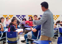 Homem de pé usando camisa de manga comprinda cinza fla para jovens que estão sentados em cadeiras escolares azuis.