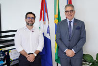 Há dois homnes posando para foto. Um veste camisa de manga longa branca e outro veste terno cinza.Ambos usam óculos.