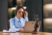 Mulher em ambiente de escritório jurídico trabalha em um notebook, aparentando concentração, enquanto ajusta os óculos. Em primeiro plano, há uma pequena estátua da Justiça (com balança e espada) sobre a mesa, reforçando o contexto profissional ligado ao direito.