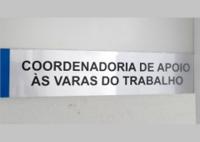 Foto de uma  placa afixada em uma porta cinza com o nome: Coordenadoria de Apoio às Varas do Trabalho escrito em letras pretas