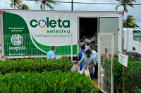 Um contêiner de coleta seletiva, identificado com a marca “Coleta seletiva”, está aberto enquanto alguns trabalhadores retiram materiais recicláveis de seu interior. A estrutura fica cercada por vegetação bem cuidada, com palmeiras ao fundo, sugerindo um ambiente externo organizado e sustentável.