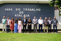 A imagem mostra um grupo de onze pessoas posando lado a lado em frente à fachada da Vara do Trabalho de Atalaia. Ao fundo, há uma parede revestida com pequenos azulejos azuis, onde se lê, em letras grandes e claras: “VARA DO TRABALHO DE ATALAIA”.  Os integrantes do grupo estão vestidos de forma formal e semiformal, com trajes que variam entre ternos, camisas sociais e roupas mais leves, sugerindo um ambiente institucional, mas também descontraído. Eles estão alinhados sobre uma área de grama.