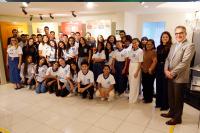 Um grupo numeroso de estudantes, acompanhado por alguns adultos, posa para uma foto em um espaço expositivo interno. Os jovens, em sua maioria usando camisetas padronizadas, estão organizados em fileiras, alguns agachados na frente e outros em pé atrás. Ao fundo, há painéis informativos sobre temas históricos, e o ambiente é bem iluminado, com aparência de museu ou memorial.