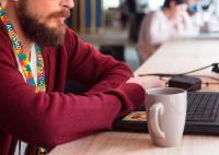 Homem com barba, vestindo casaco vermelho, trabalha em um notebook sobre a mesa, ao lado de uma caneca. Ele usa um cordão colorido com a inscrição “autismo – conscientização”, enquanto o ambiente ao fundo aparece desfocado, sugerindo um espaço de trabalho compartilhado.