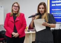 Há duas mulheres de peles claras. A primeira veste um terno vermelho e calça preta. Já a segunda, veste um vestido escuro e está segurando um microfone e um papel. 