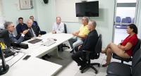 Grupo de pessoas participa de uma reunião em uma sala administrativa do TRT-AL, com integrantes sentados ao redor de uma mesa enquanto um homem de terno conduz a conversa. O ambiente conta com computadores, televisão e cadeiras distribuídas pelo espaço.