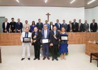 A imagem mostra uma solenidade realizada em um plenário, reunindo autoridades, homenageados e convidados em um registro oficial coletivo. Na parte frontal, cinco pessoas posam segurando placas comemorativas e medalhas recebidas durante a cerimônia. Ao centro, destaca-se um grupo de autoridades e representantes institucionais posicionados diante da mesa principal do espaço. O ambiente possui acabamento em madeira clara, iluminação moderna e um crucifixo fixado na parede ao fundo. A fotografia transmite um cl