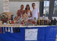Uma banca de feira com três pessoas sorrindo atrás de uma mesa cheia de produtos artesanais. Sobre a mesa há velas, colares, frascos com líquidos e itens naturais organizados. Ao fundo, há uma parede com azulejos claros e cartazes. O ambiente parece um mercado ou evento ao ar livre.