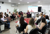 Grupo de pessoas sentadas em cadeiras pretas. Há um tapete vermelho no centro da sala. As pessoas ouvem uma mulher que está de pé com um microfone na mão.
