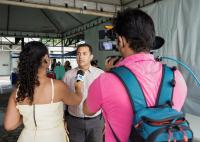 Há um homem de camisa clara falando ao microfone. Ele está sendo entrevistado por repórtes de vestido claro. O cinegrafista está com cmisa rosa e tem uma mochila azul nas costas. Ele segura uma câmera.