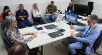 A imagem mostra uma reunião realizada em um ambiente de escritório, com a participação de magistrados, servidores e jornalistas. Sentados ao redor de uma mesa, os participantes acompanham atentamente a fala de um homem de terno azul, que conduz a conversa. Sobre a mesa, há notebooks, canetas e documentos utilizados durante o encontro. Ao fundo, computadores e equipamentos reforçam o ambiente institucional e de trabalho.