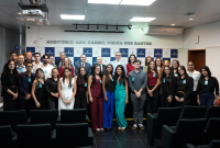 Voto de várias pessoas em pé posadas para a foto em um auditório solene, entre homens e mulheres. Ao fundo, quatro homnes utilizando ternos