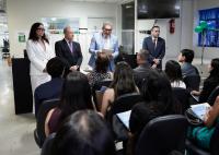 A fotografia mostra uma solenidade institucional realizada em um ambiente interno do TRT-AL, com autoridades posicionadas à frente do público. Ao centro, um magistrado faz uso da palavra enquanto segura um documento, acompanhado de outros representantes da instituição. Na plateia, servidores e participantes acompanham atentamente a cerimônia sentados em cadeiras organizadas em fileiras. O espaço está decorado com balões e elementos nas cores azul e verde, reforçando o caráter comemorativo do evento.