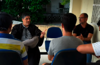 A foto mostra um grupo de trabalhadores reunidos em um ambiente interno, durante uma conversa conduzida pelo juiz Cláudio Márcio Lima Santos. Sentado à frente dos participantes, o magistrado gesticula enquanto dialoga com os presentes, em um momento de escuta e orientação.