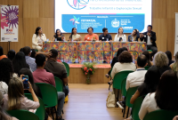 A imagem mostra um auditório durante um evento promovido pelo Fórum Estadual de Prevenção e Erradicação do Trabalho Infantil e Proteção ao Adolescente Trabalhador de Alagoas (FETIPAT/AL). Na mesa principal, sete participantes acompanham a fala de um dos debatedores, enquanto o público assiste atentamente. O espaço é decorado com elementos coloridos e um painel ao fundo destaca o tema relacionado ao enfrentamento das violências, do trabalho infantil e da exploração sexual.
