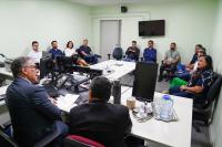 A imagem mostra uma reunião realizada em uma sala administrativa, com cerca de uma dúzia de participantes sentados ao redor de uma mesa e ao longo das paredes. Em primeiro plano, dois homens de terno conduzem ou acompanham a discussão, enquanto os demais observam atentamente. O ambiente conta com computadores, telefone, documentos e equipamentos de escritório, indicando uma atividade de trabalho ou visita institucional. Os participantes demonstram atenção às falas apresentadas durante o encontro. 