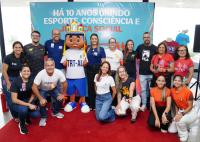 Grupo de pessoas reunidas em volta de um mascote que está com uma camiseta branca escrito TRT-AL e uma bermuda azul e calça tênis branco.