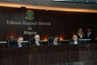 A imagem mostra uma sessão solene no plenário do Tribunal Regional Eleitoral de Alagoas (TRE/AL), com magistrados e autoridades posicionados à mesa de honra, tendo ao fundo a identificação institucional do tribunal. Ao lado direito, duas pessoas acompanham a cerimônia em pé.