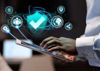 A imagem mostra uma pessoa utilizando um tablet enquanto interage com uma interface digital representada por elementos gráficos em tons de azul. No centro da composição, destaca-se um escudo com um símbolo de verificação, remetendo à segurança e à proteção de dados. Ao redor dele, aparecem ícones que representam documentos, usuários, configurações, certificação, compartilhamento de informações e conexão global, sugerindo conceitos relacionados à segurança cibernética, governança digital.