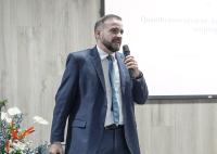 Há um homem de terno falando ao microfone. Há um quadro branco atrás dele e um arranjo de flores